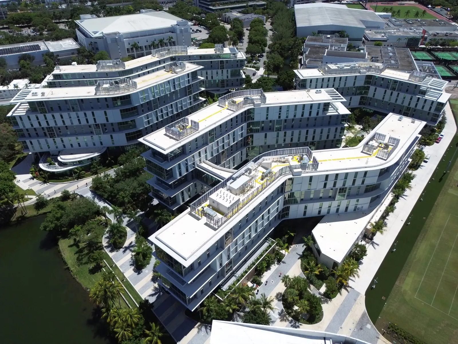 An aerial view of curving white residential buildings beside a lake, with palms, walkways and playing fields around them